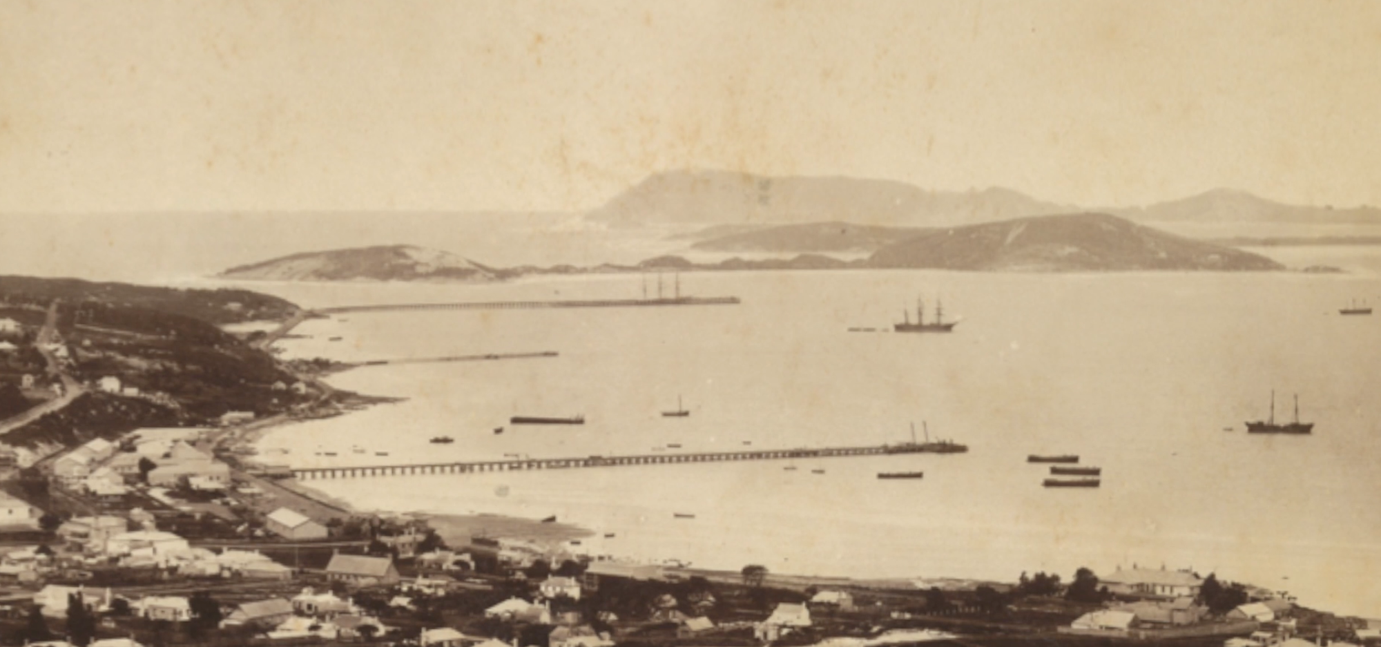 Sepia-toned panoramic view of a coastal town and harbour, with long jetties, anchored ships, and distant hills across the water.
