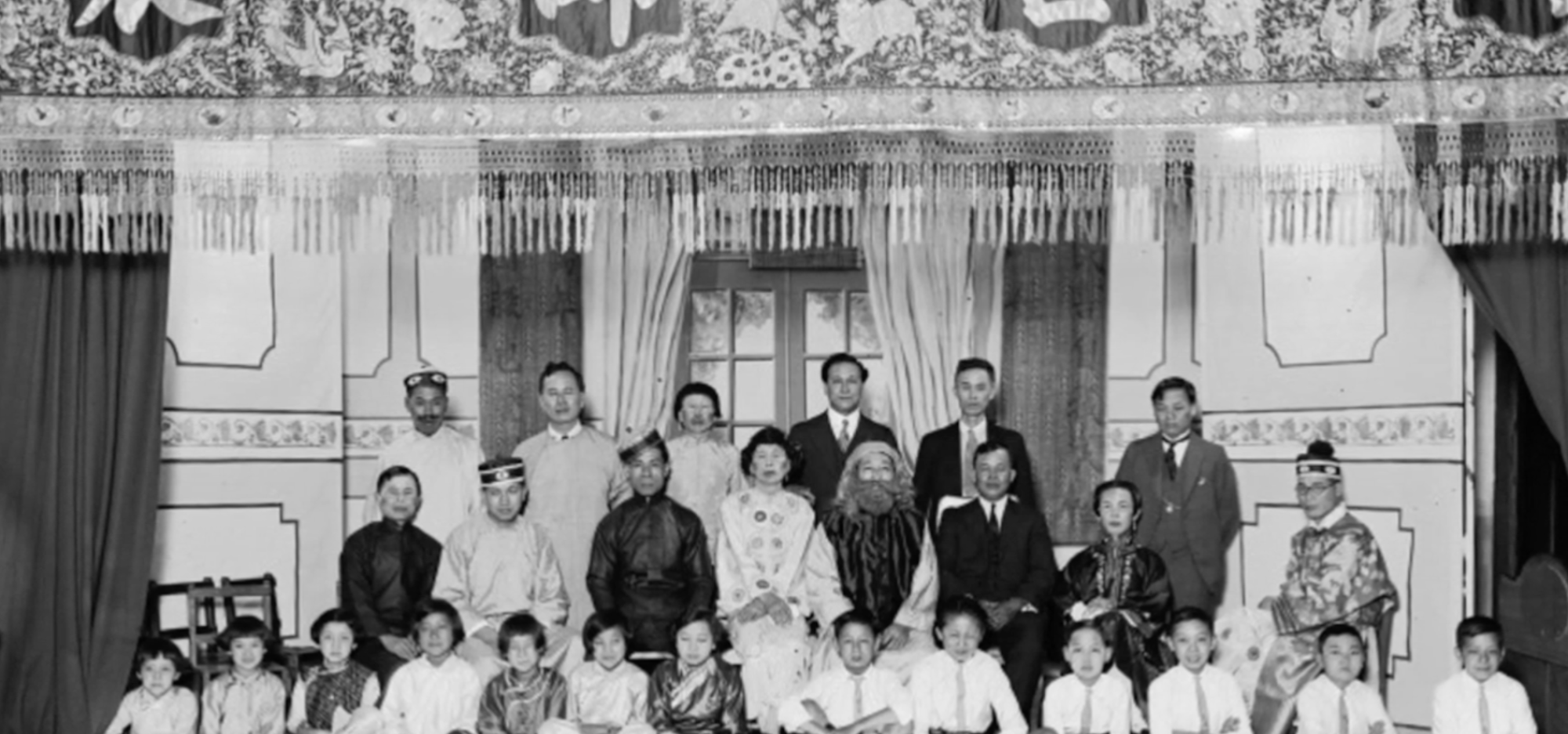 Black-and-white group portrait of adults and children posed in rows inside an ornate interior with decorative curtains and wall panels.