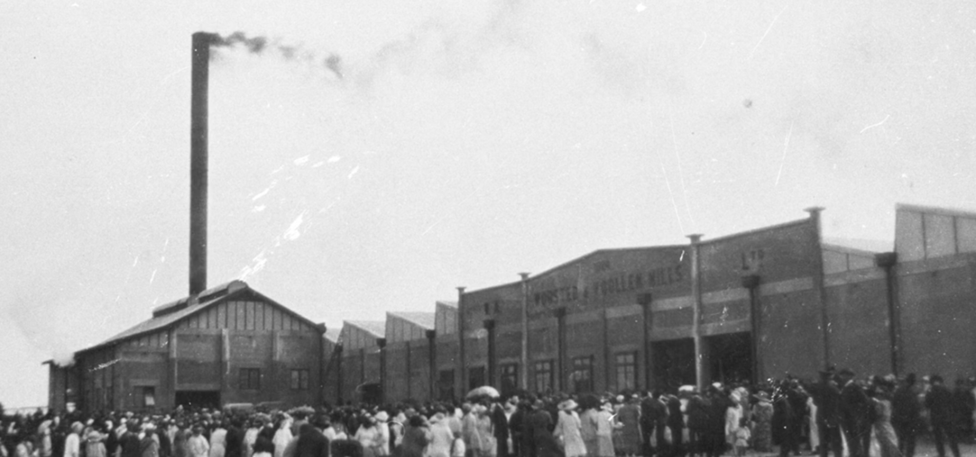 Black-and-white photo of a large factory with a tall smokestack, where a crowd of people is gathered outside the building.