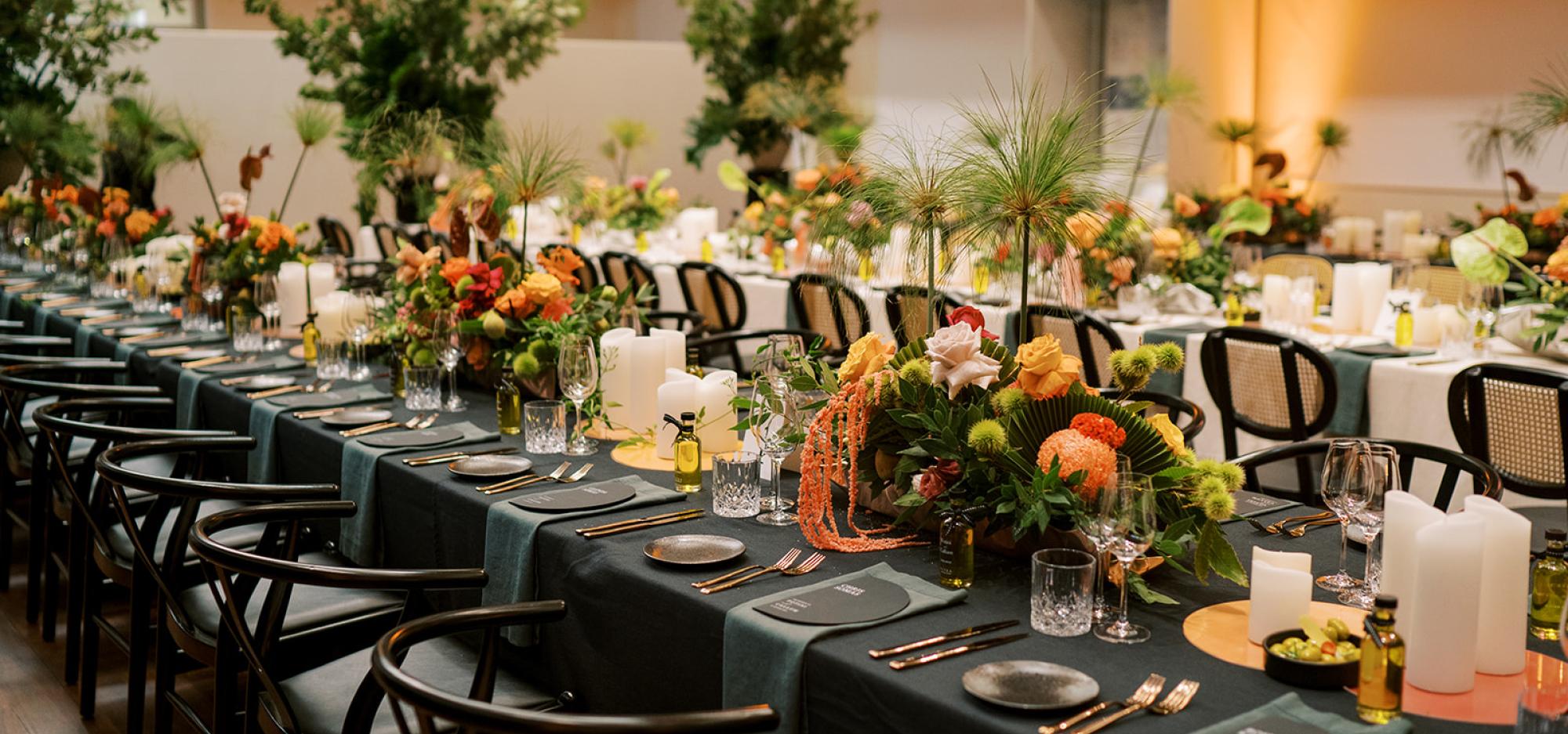 A formal dining area set up with tables covered in black table cloths, floral arrangements and greenery.