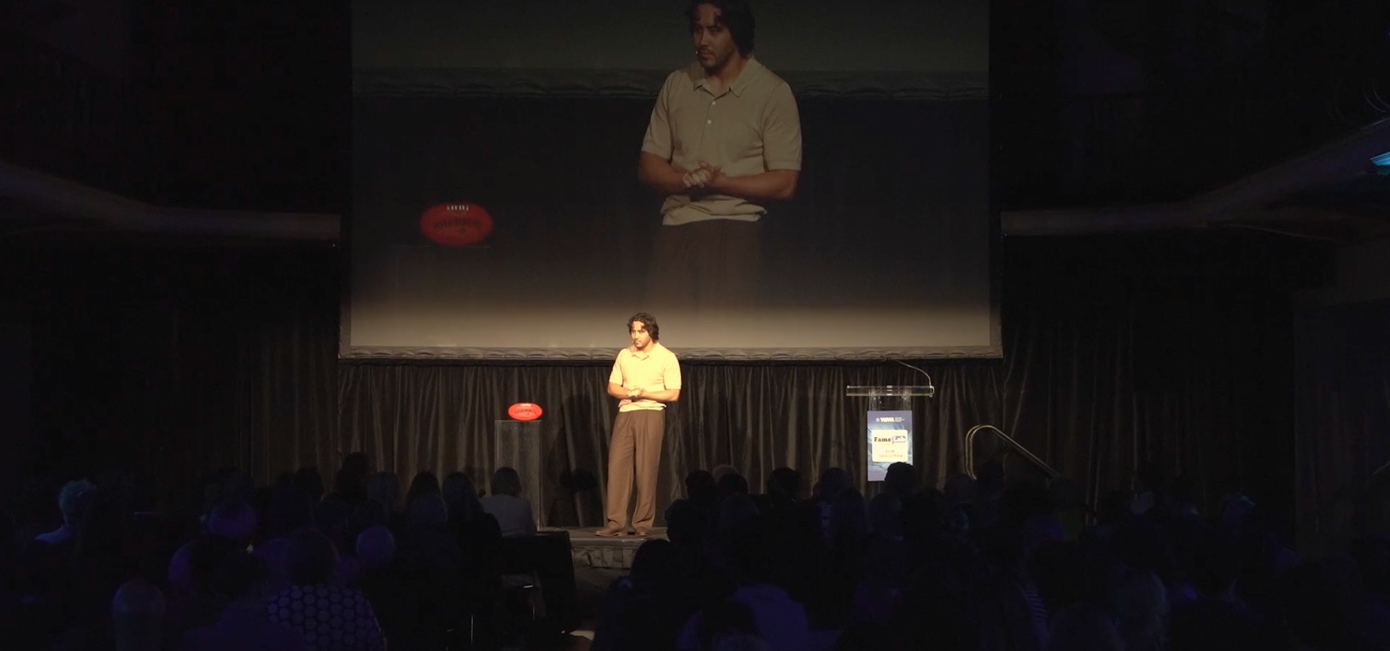 Presenter standing on a stage in front of a seated audience, with a large screen behind displaying a live close-up of the speaker.