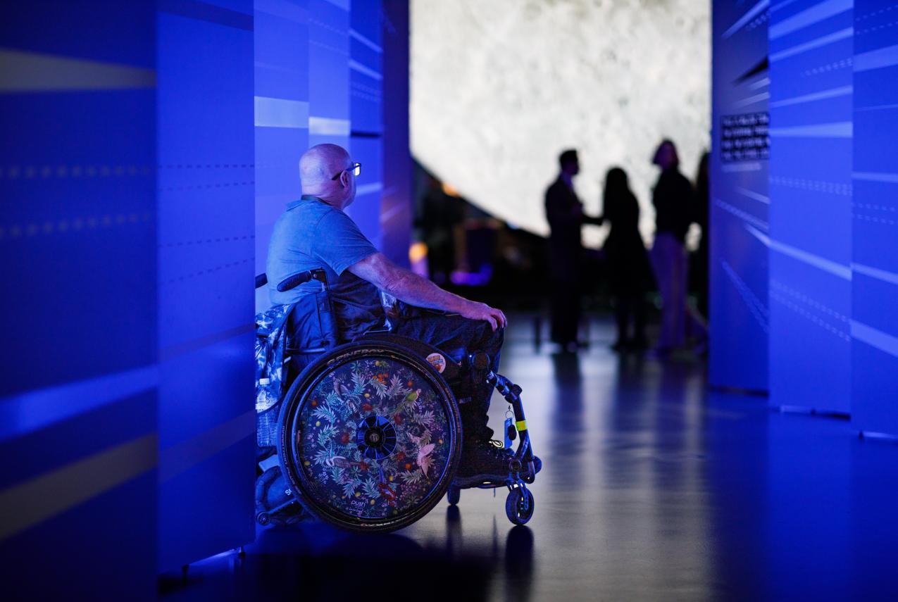A person in a grey shirt and black glasses with a bald head sits in a wheel chair with an intricately decorated wheel featuring drawings of Australian birds and foliage. They are seated in a vividly-lit blue hallway with a futuristic design in front of a large moon sculpture
