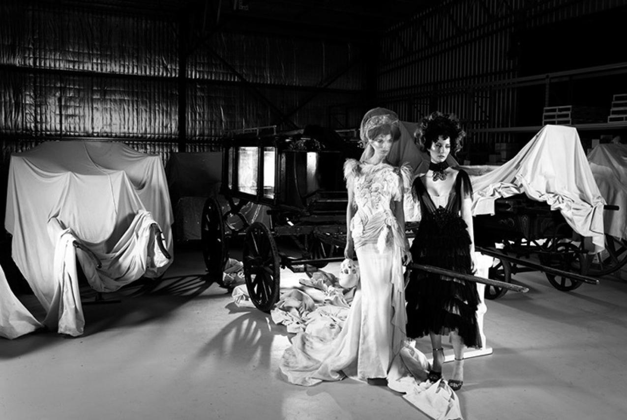 Two models in elaborate gowns stand in front of partially covered carriages in a dimly lit storage space.
