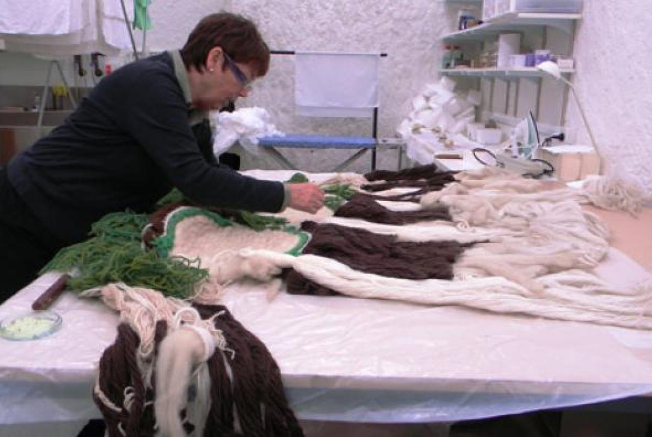 Person working on a textile project with yarn strands of green, brown, and cream on a table in a craft workspace.