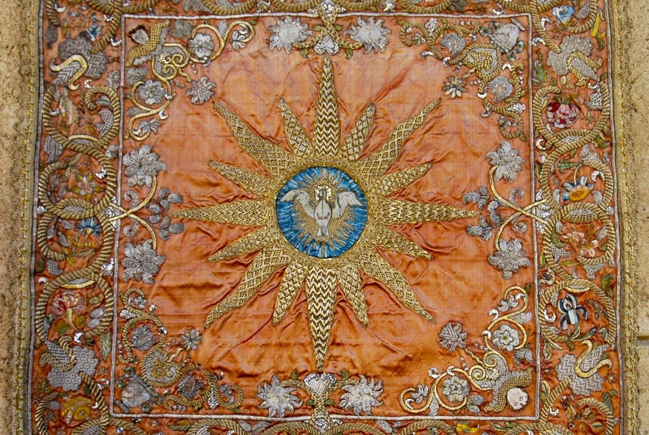 An embroidered textile with a central pelican emblem and sunburst on an orange background, surrounded by floral motifs.