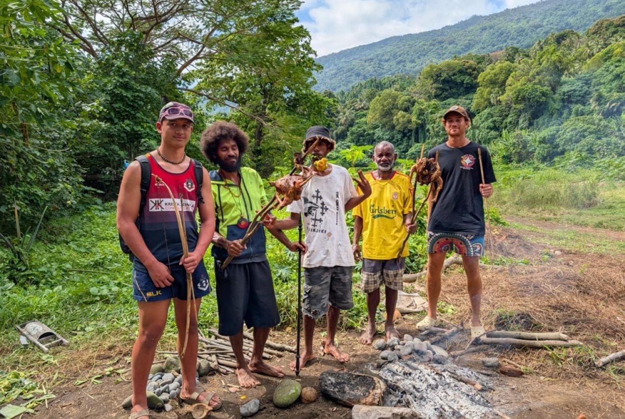 Five Indigenous young people standing around a small fire holding sticks with meat while surrounded by greenery