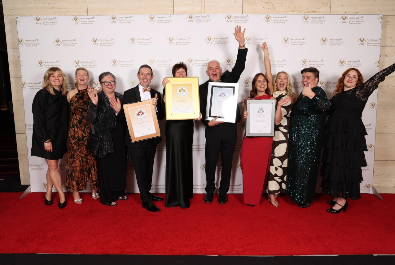 A happy group of 10 people from the WA Museum holding award certificates Infront of the Tourism award media wall after receiving an award