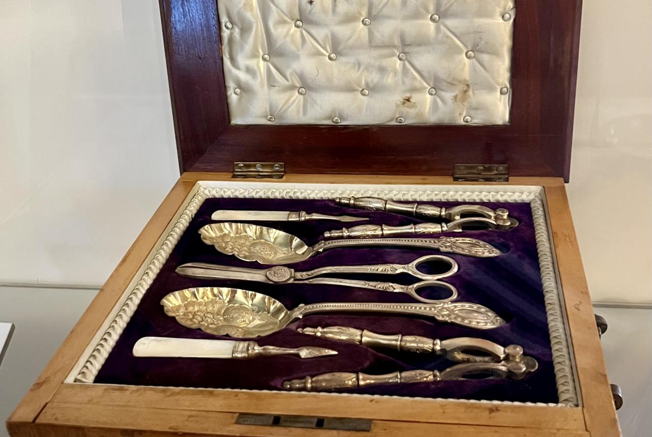 An open wooden presentation box with a satin-padded lid and a deep purple velvet interior, displaying an ornate Victorian-era silver serving utensil set. The set includes intricately designed spoons, tongs, and scissors with decorative handles, arranged neatly inside the box.