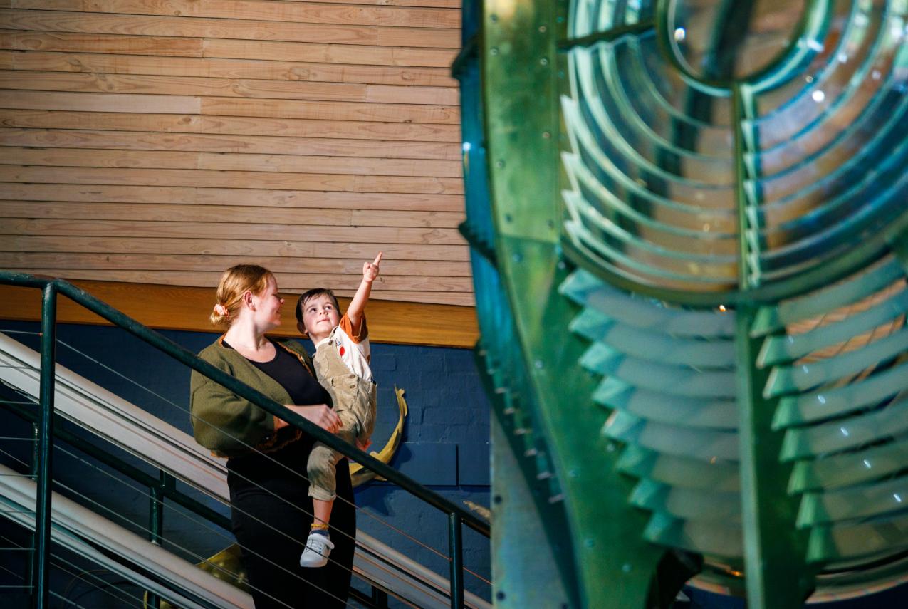 A woman carries a small child up a staircase as the child excitedly points toward a large, illuminated museum exhibit.