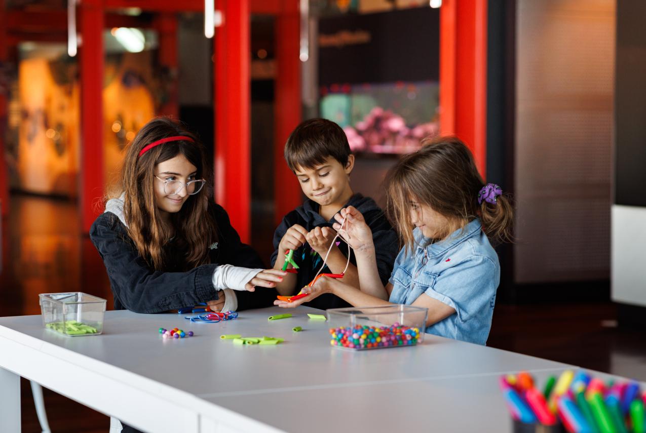 Three children seated at a white table in a museum activity area, working on a craft project with colorful beads, pipe cleaners, and other art supplies. Clear containers of materials are on the table, and bright red structural elements and exhibit displays are visible in the background.