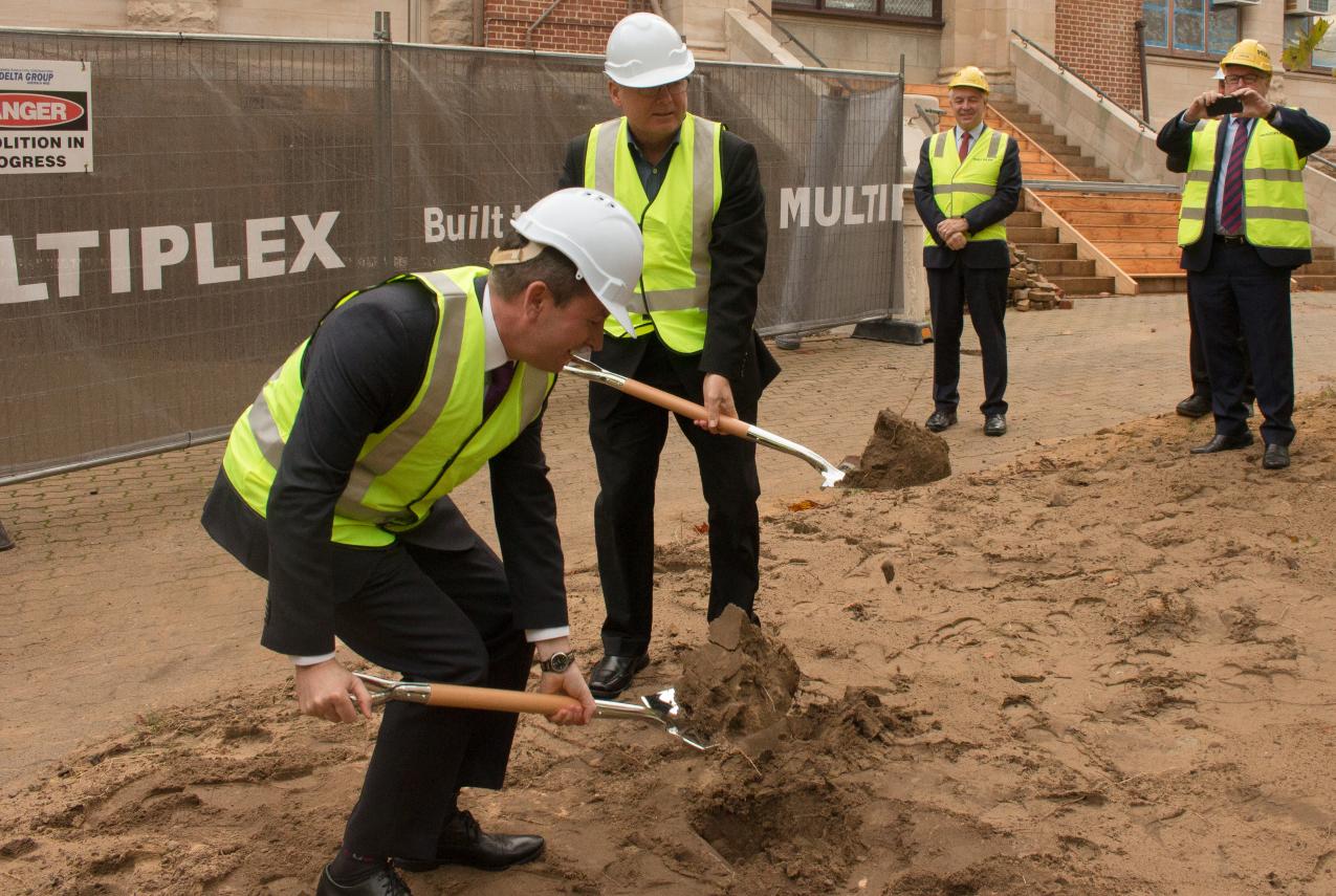 Premier McGowan bending with a ceremonial shovel and Minister Templeman standing to his left.  They are turning sod by digging the soil at the site of the New Museum.  Both are wearing fluro yellow vests and hard hats.