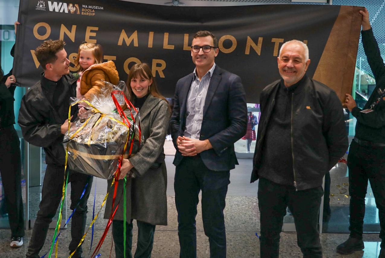 Four adults and a toddler stand in front of a banner that says "One Millionth Visitor" held by two Museum staff members.