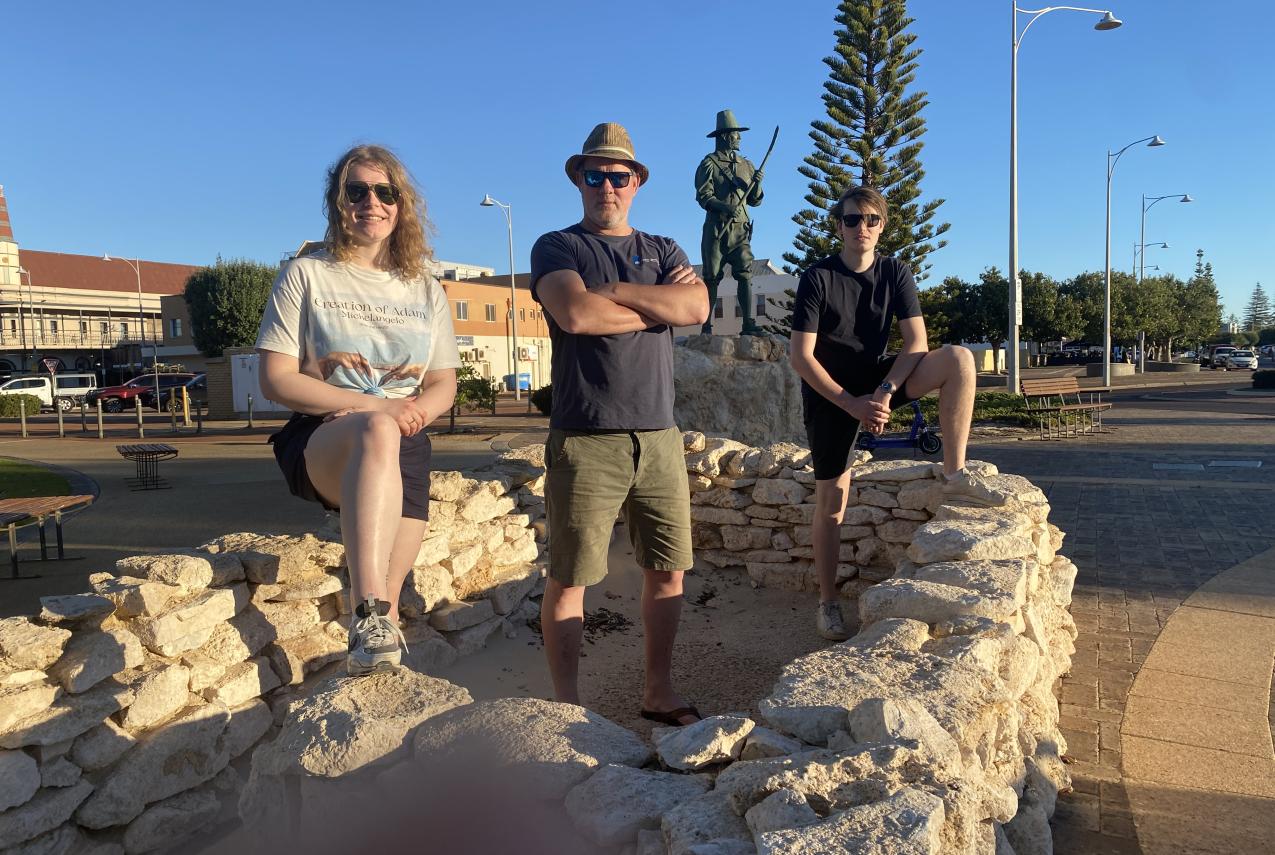Three people stand on a circular stone structure with a statue and a tree in the background.