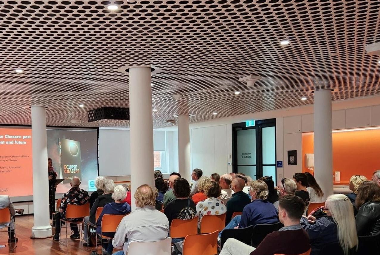 A large group of people seated in orange and white chairs face a large projector at the front of the room with an individual standing at a podium. The image projected onto the screen displays a book titled 'Eclipse Chasers' and some smaller text