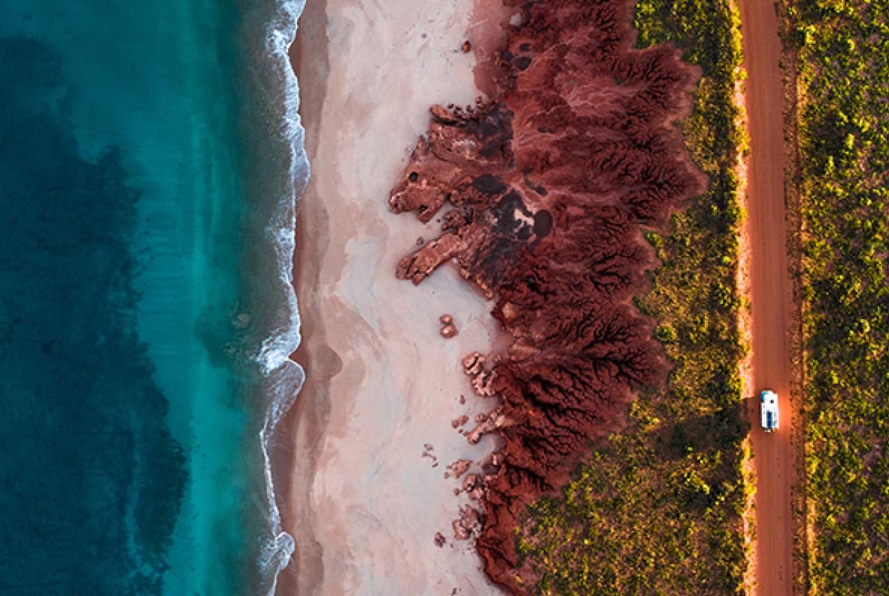 Ocean, red dirt and car driving along a dusty road - aerial shot.