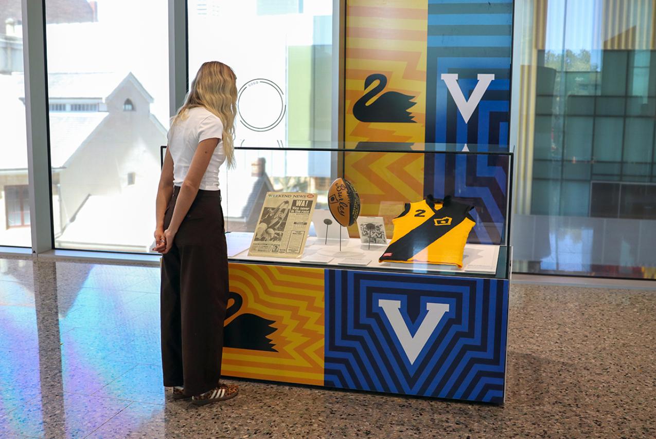 A woman looks at a small showcase of AFL memorabilia in a Museum. The showcase is surrounded by the WA and Victorian Football logos.