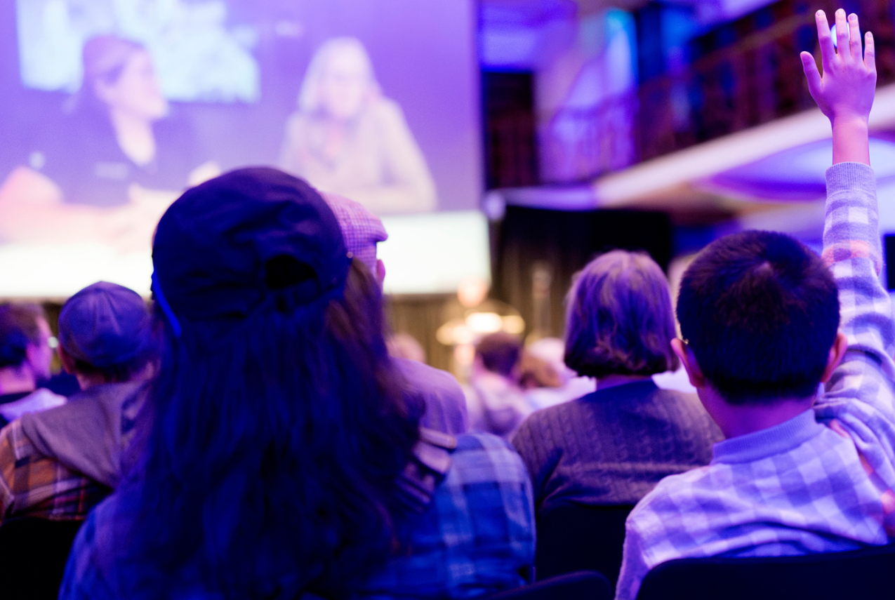 Audience members seated at a conference or presentation, with one attendee raising a hand to ask a question while speakers appear on a large screen at the front of the room.