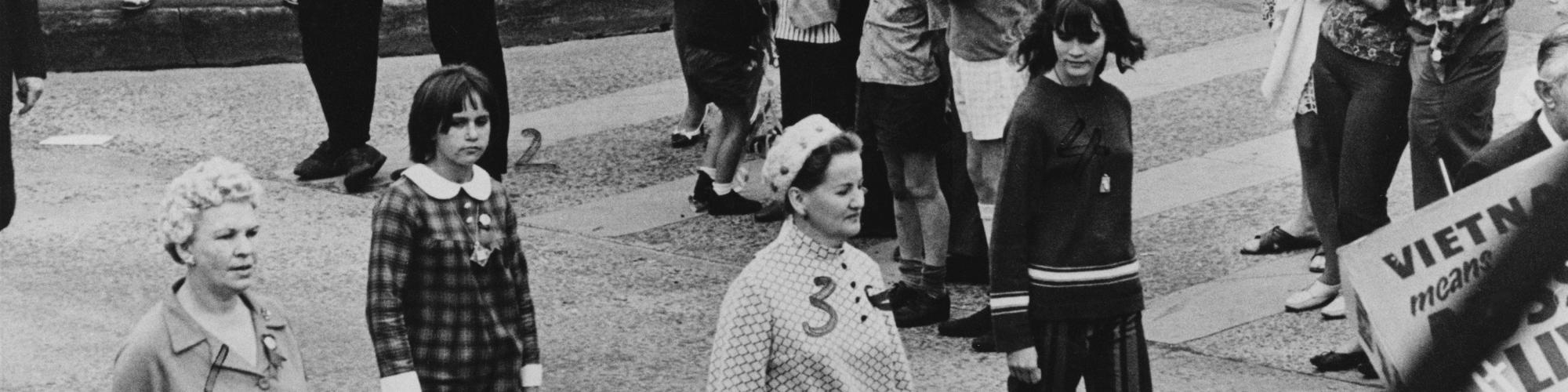 A black and white photograph of people dressed in 1960s-style clothing walking on a street