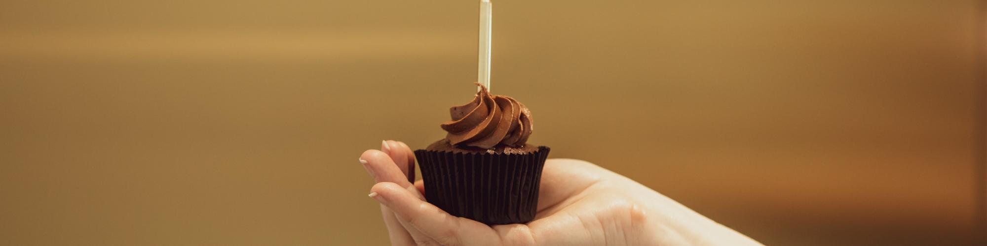 A hand holding a chocolate cupcake with a golden birthday candle