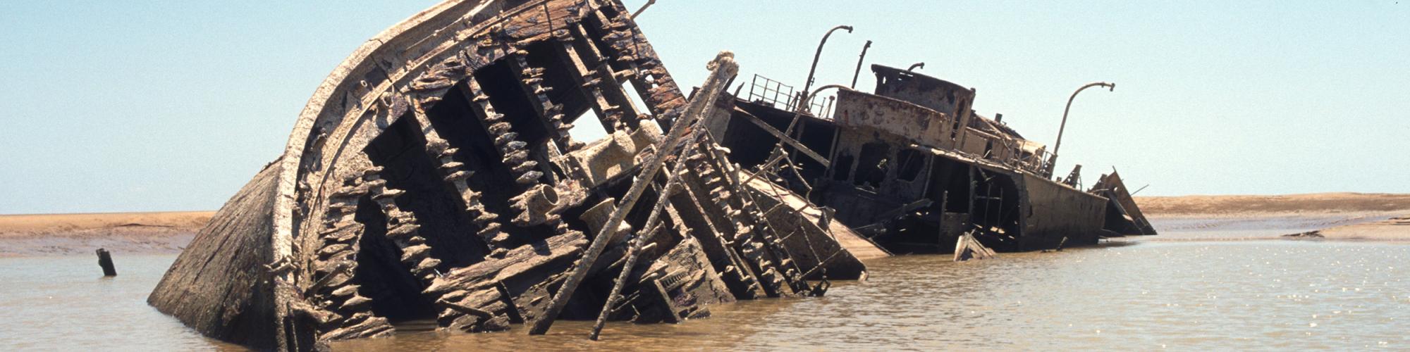 A shipwreck in shallow water near a sandbar, beneath a light blue sky. Most of the upper half of the shipwreck is exposed to the air above the water's surface.