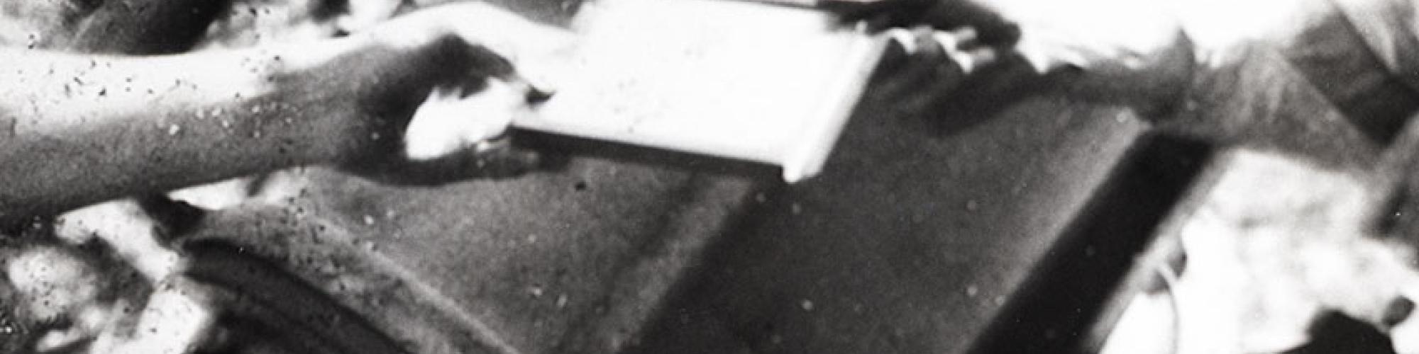 black and white image showing two hands holding a photographic slide. one hand on the right and other on left of photograph edge to indicate they belong to different people and are passing the slide to one another. A large drum of a telescope with viewing device is in the background. 