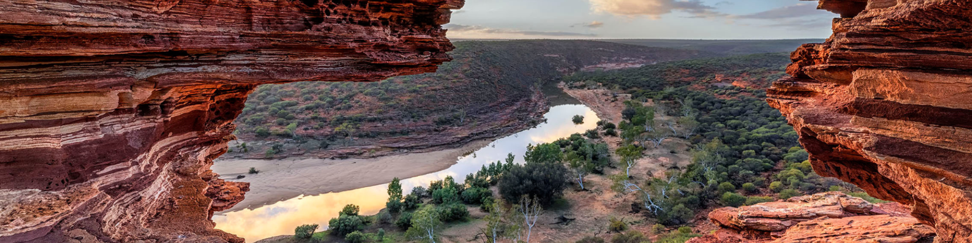 A photograph of Nature's Window, a rock-formation in Kalbarri, with the camera peeing through a colourful banded rock hole looking out over a green and brown landscape and blue sky