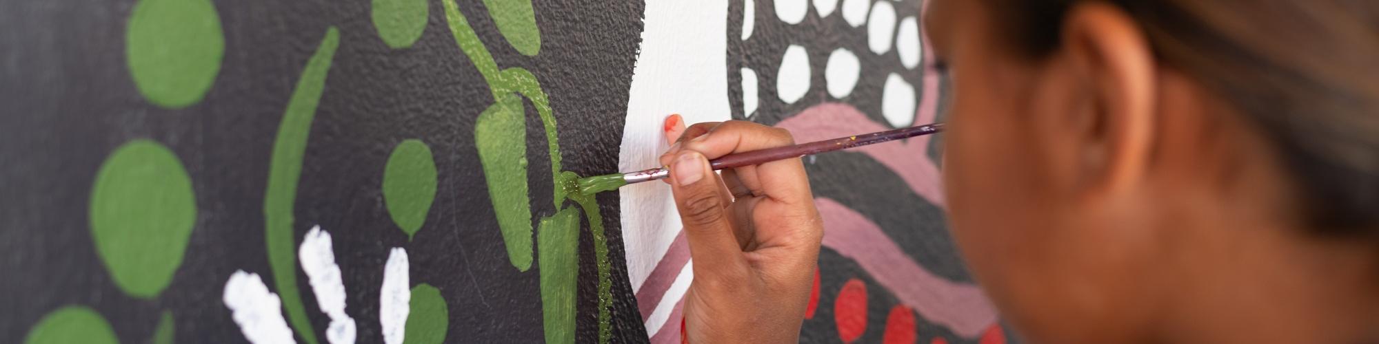 A person with brown hair paints a mural with prodominantly black, green, yellow colours. you cant see the persons face but from behind them as the mural takes shape