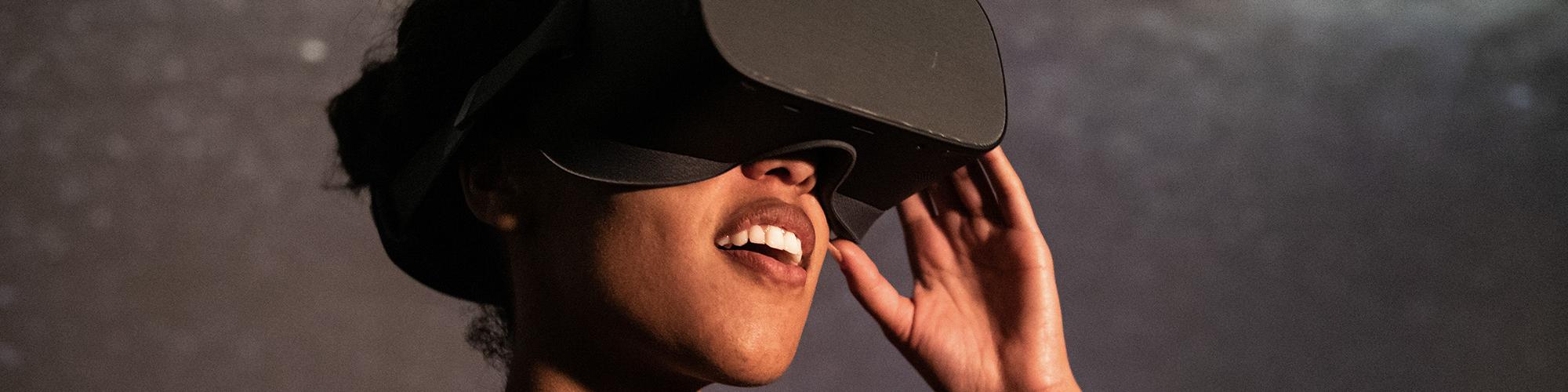 A person wearing a black button up shirt wears a black VR set and smiles as though out of wonder and enjoyment as they look through the headset. They stand in front of a galaxy backdrop