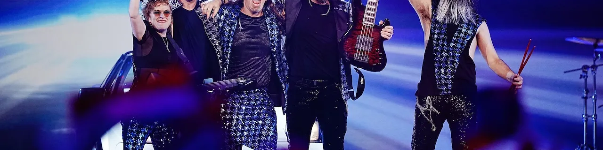 The five members of Voyager dressed in matching silver and black rockstar ensembles cheer and celebrate on the brightly lit Eurovision stage. 