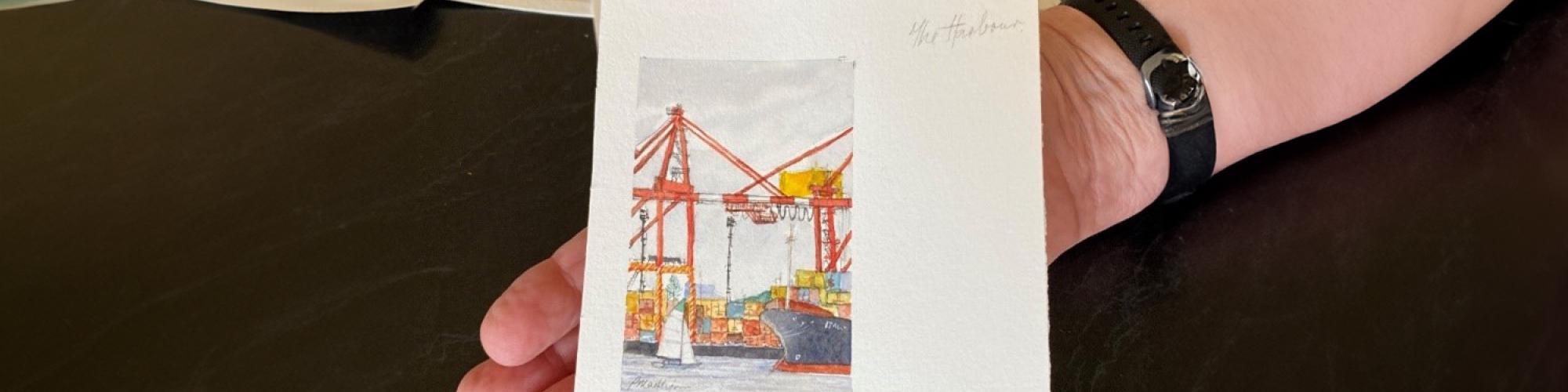a small painting of a scene in Fremantle is being held in the palm of a persons hand. Other paintings are in the background