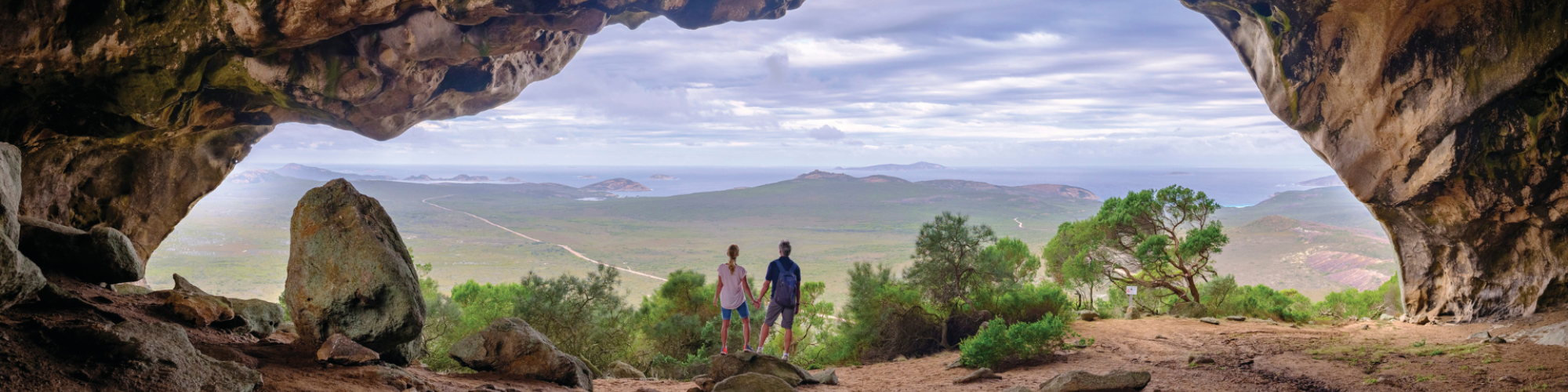 A wide angled shot of two people holding hands with their back to the camera, standing in the mouth of a large cavern. A green landscape stretches out before and beneath them