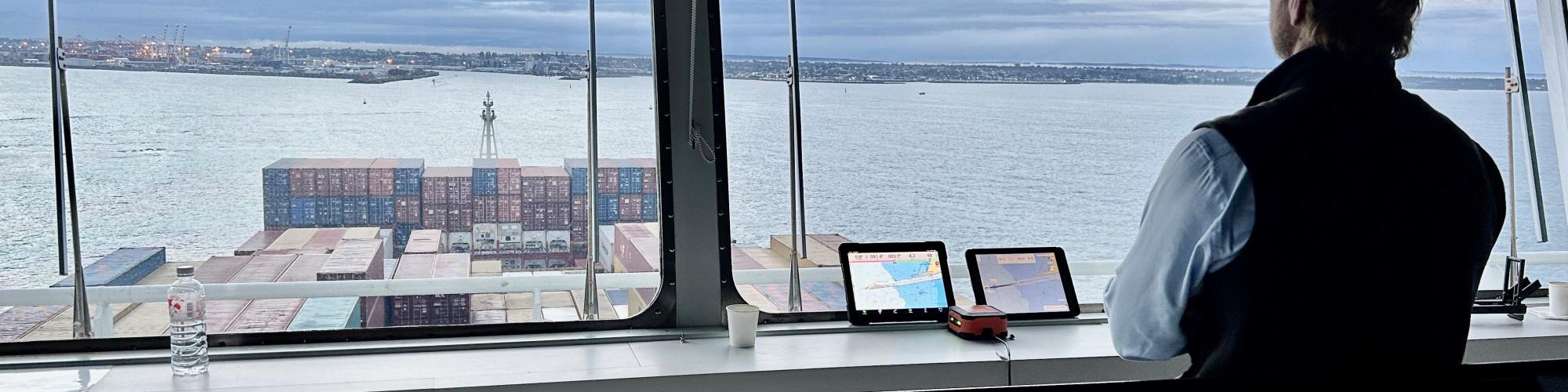 an adult person in slight silhouette stands inside a ship indoor viewing deck with controls. They look out through the large window onto a harbour with sea containers