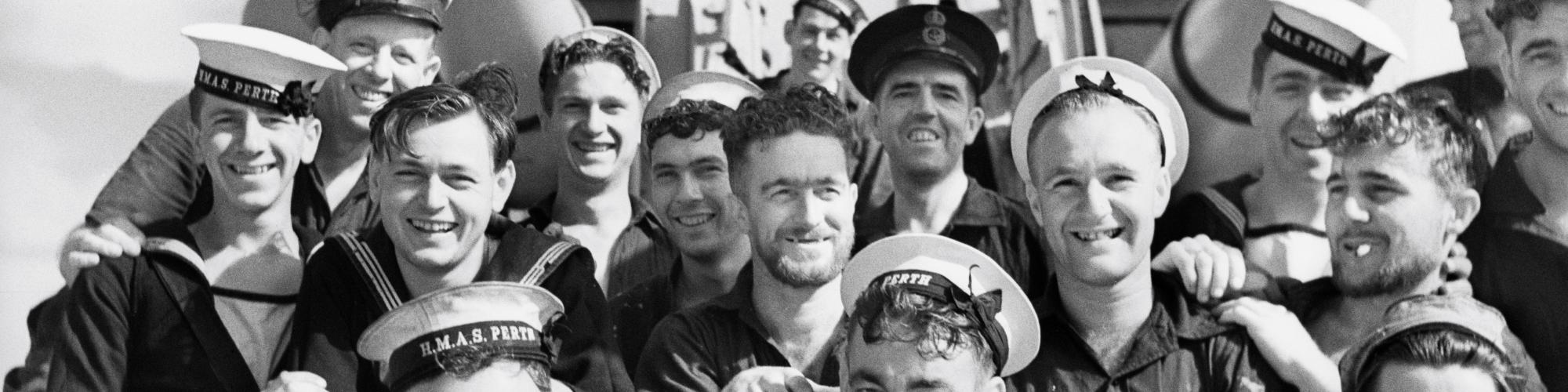 group of men in navy uniform on HMAS Perth smiling for a photo sitting under the ships guns