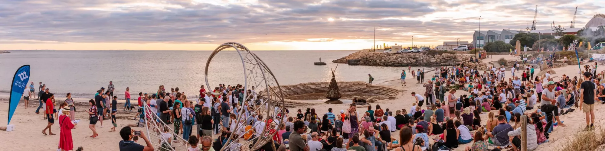 A panoramic shot of a crowded beach at early dusk facing towards a purple and grey ocean with golden highlights. People sit around decorative sculptures on the sand.