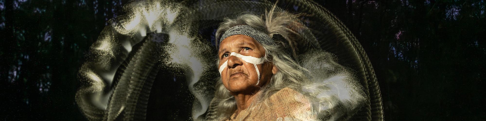 an Aboriginal Elder with traditional ochre on her face and arms looks up. She wears clothing made of emu feathers. There is a white circular halo and dust around her