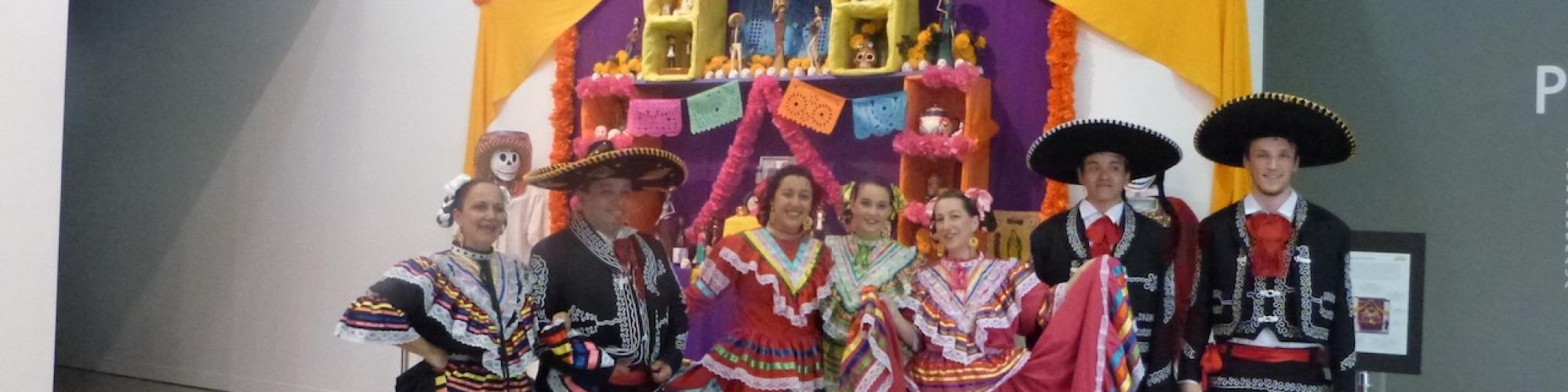 A group of people in colourful traditional costumes standing in front of a decorated Día de los Muertos altar inside a gallery space.