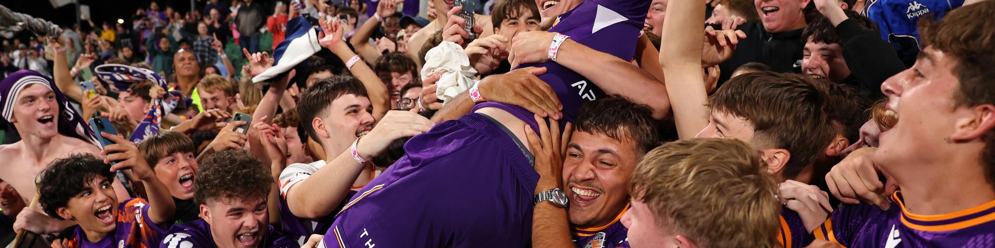 Perth Glory player celebrates with supporters in the stands after a match, surrounded by a cheering crowd in club colours.