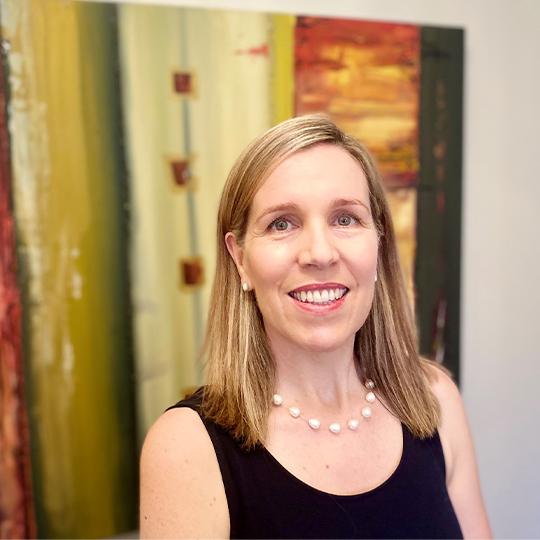 A woman with long blonde hair stands in front of an artwork, smiling at the camera