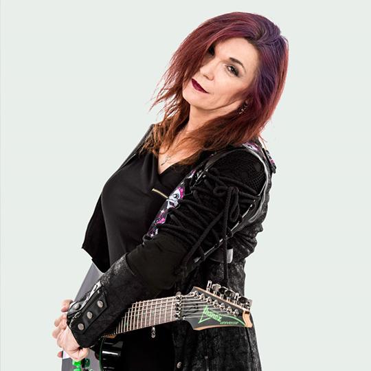 A woman with dark red hair wearing a long black coat stands holding an electric guitar