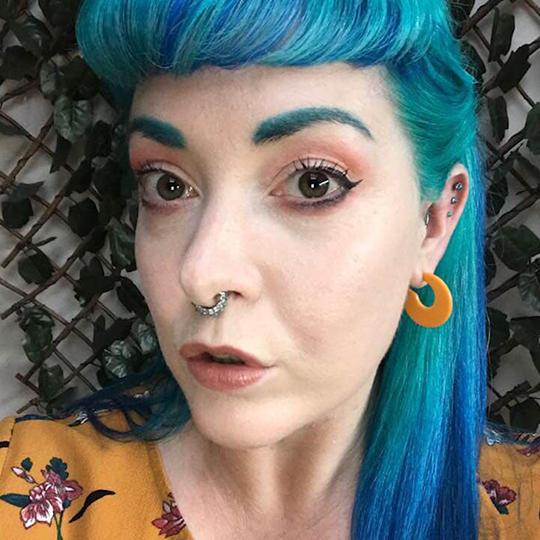 A young woman with vibrant blue hair stands in front of a plant trellis, looking seriously at the camera