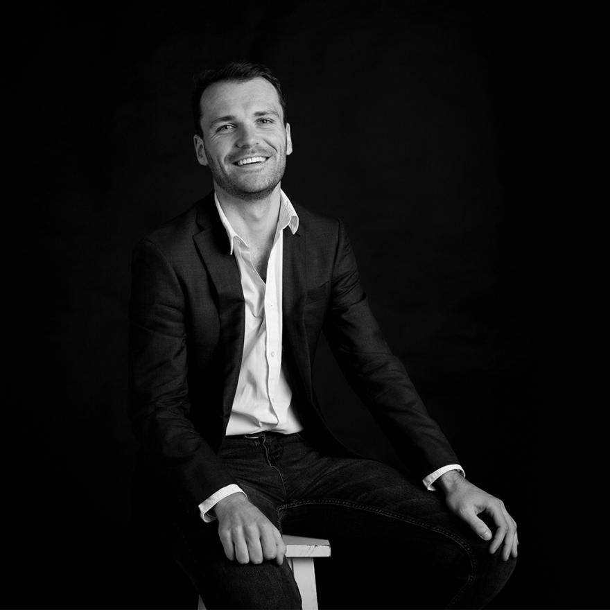 A studio photograph of a young man wearing a suit jacket and jeans, smiling at the camera