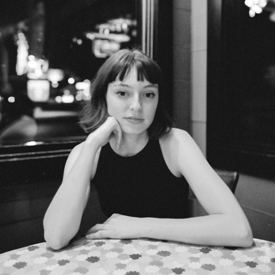 A young woman with short dark hair sits at a red-lit corner booth in a cafe, looking seriously at the camera