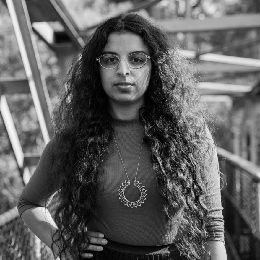 A young woman with long curly hair stands on a bridge, looking seriously at the camera