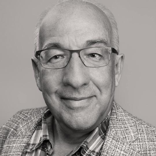 A man wearing a patterned shirt and glasses smiles at the camera