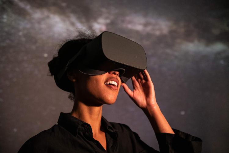 A woman wearing a VR headset stands in front of a starry projected backdrop with an expression of wonder