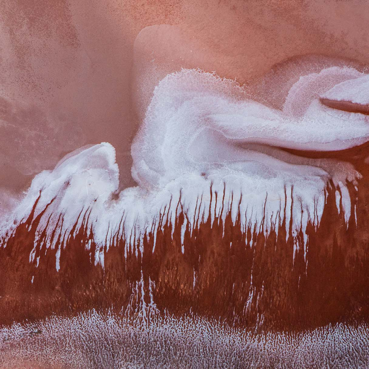 An aerial view of a hilly striated salt lake landscape in shades of red and white