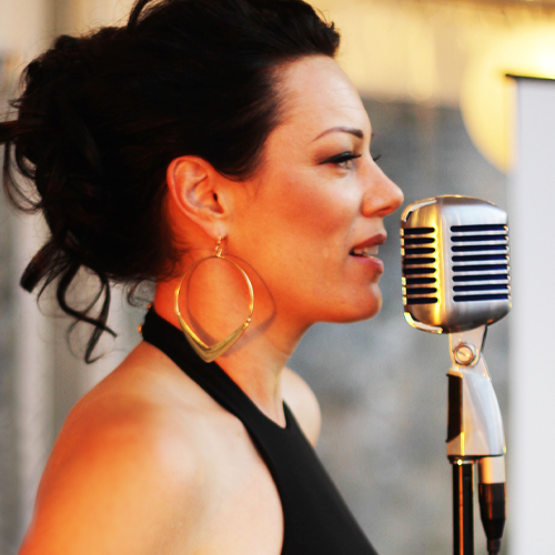 A woman wearing a black dress and large gold earrings stands in front of a retro microphone