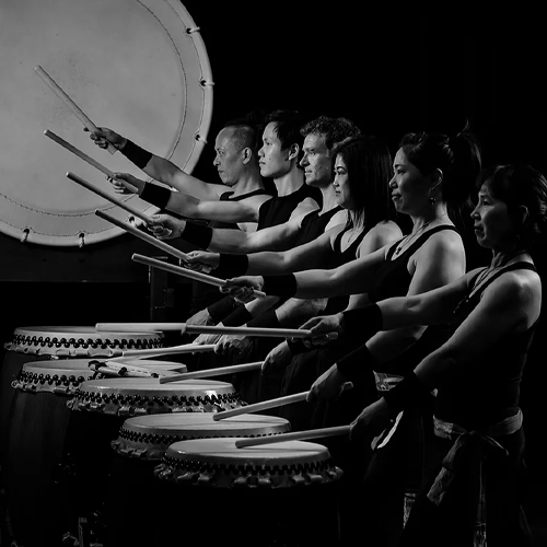 A group of Taiko drummers stand in front of their drums, each holding one drumstick aloft