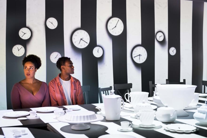 Two people with expressions of wonder sit at a outsized black and white striped table set for afternoon tea