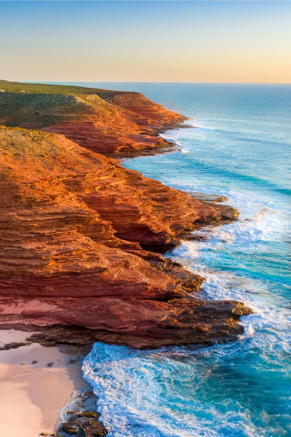 An aerial photograph of sunlit red coastal cliffs rising above the bright blue sea